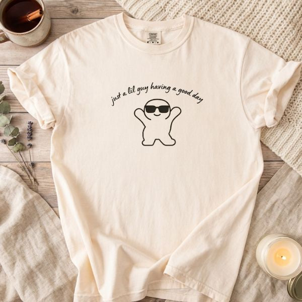 Cozy flat lay of an ivory Comfort Colors t-shirt with a "just a lil guy having a good day" graphic, styled on rustic wood with a knit blanket, a warm candle, and a ceramic mug.