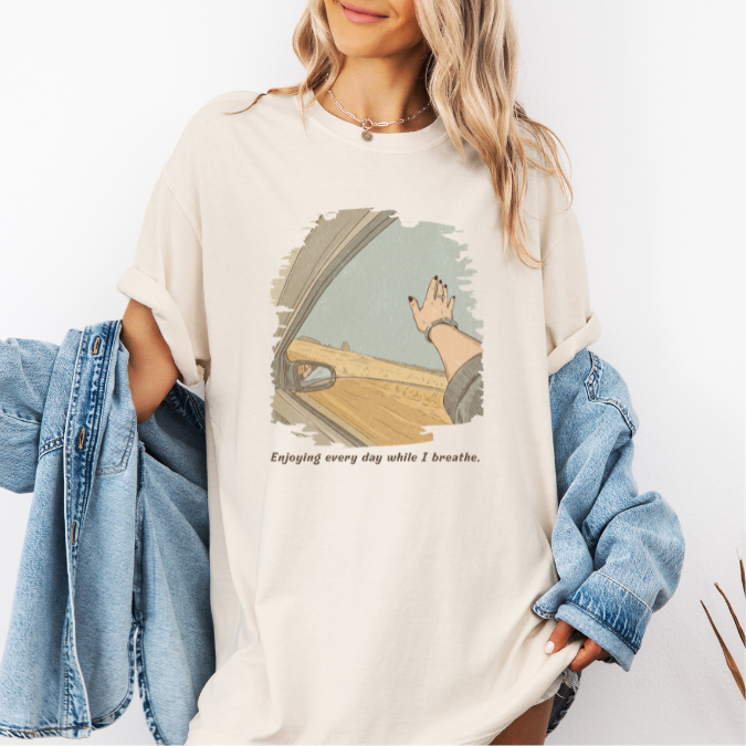 Ivory Comfort Colors graphic tee with a vintage-style open-road illustration seen from a car window and the phrase “Enjoying every day while I breathe.”