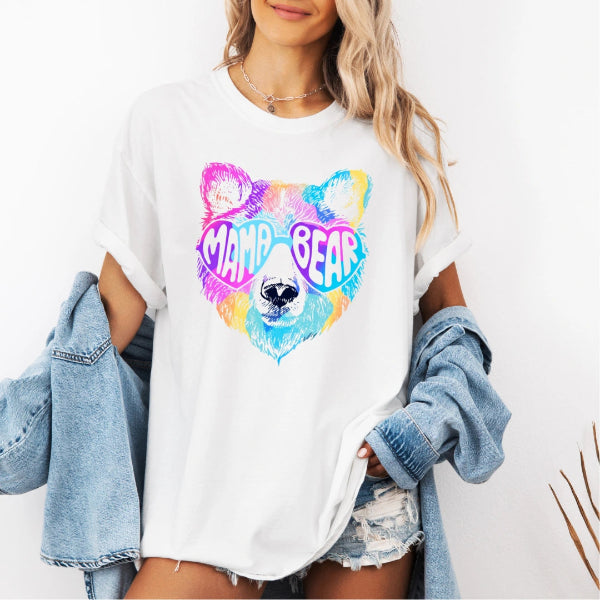 White Comfort Colors graphic tee with a colorful Mama Bear face wearing heart-shaped sunglasses and retro lettering.