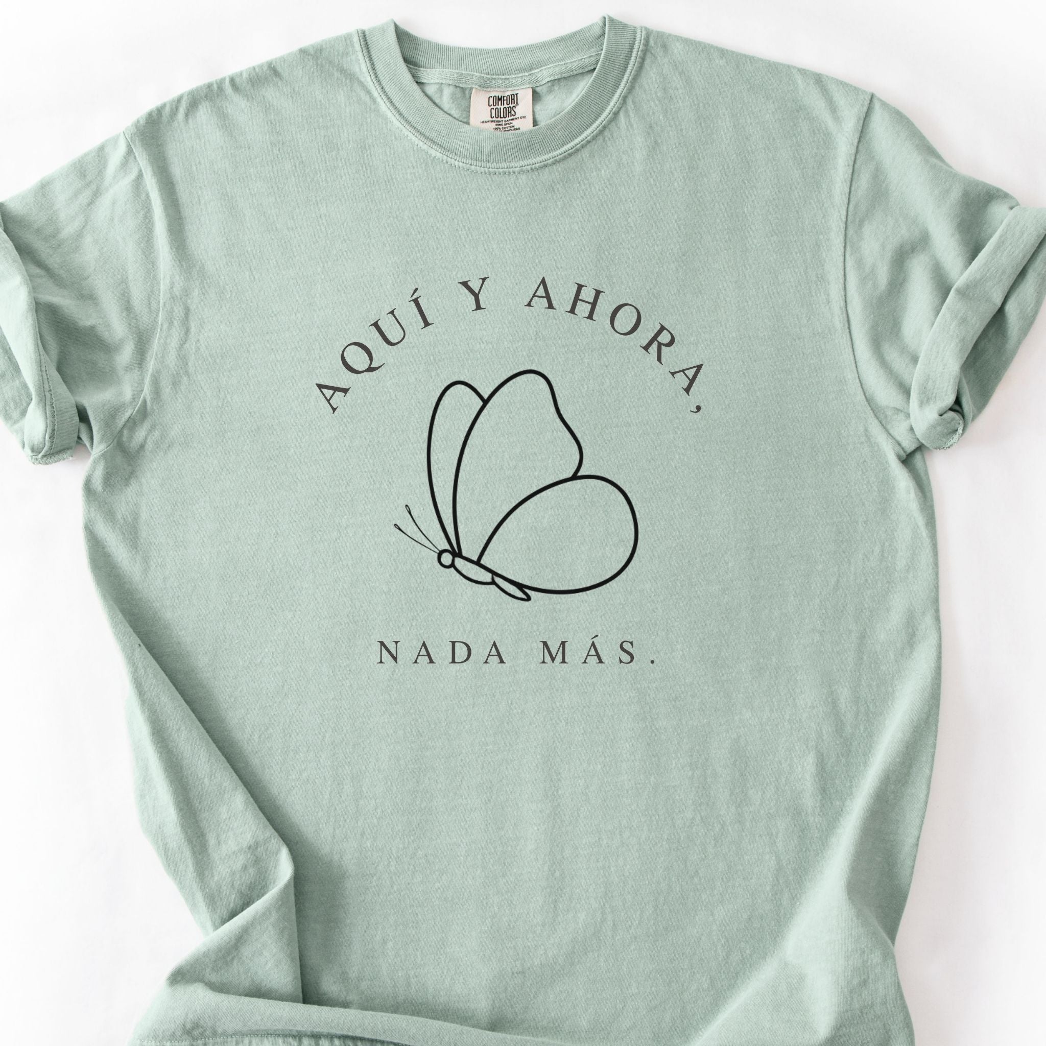 Close-up detail of Aquí y Ahora Nada Más Spanish butterfly minimalist line art printed on Comfort Colors sage t-shirt