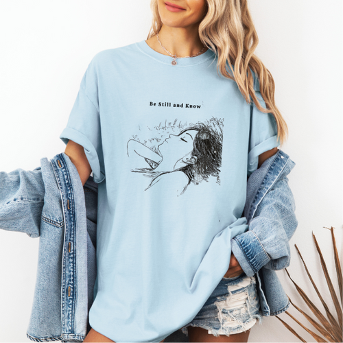 Light blue Comfort Colors graphic tee with black line art illustration of a resting woman and the phrase “Be Still and Know”