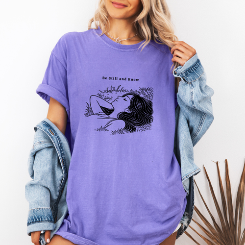 Violet Comfort Colors graphic tee with black line art illustration of a resting woman and the phrase “Be Still and Know”