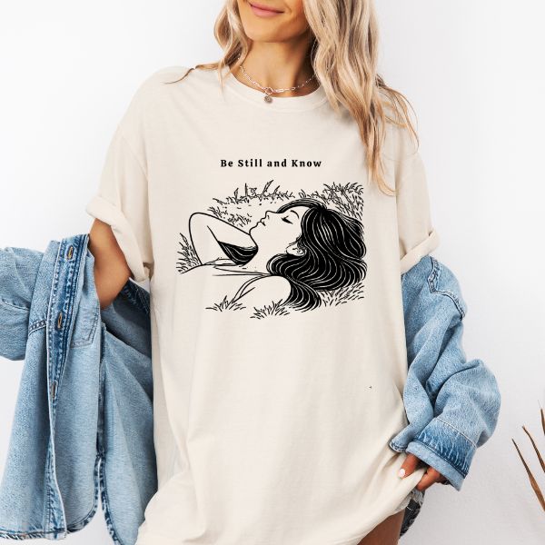 Woman wearing a natural Comfort Colors t-shirt with Be Still and Know illustration of a woman lying in tall grass with eyes closed, styled with a denim jacket