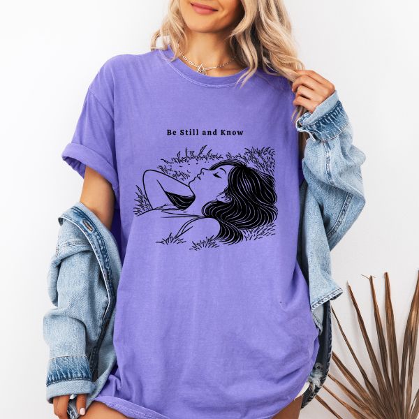Woman wearing a violet Comfort Colors t-shirt with Be Still and Know line art illustration of a woman resting peacefully in tall grass, styled with a denim jacket
