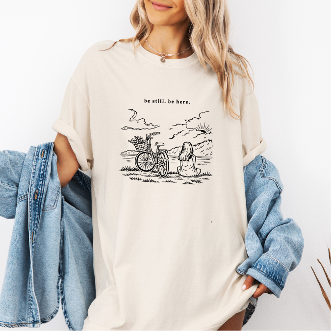 Ivory Comfort Colors graphic tee with a line art illustration of a bicycle, flowers, and a seated woman in nature with the phrase “be still. be here