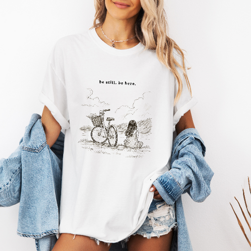 White Comfort Colors graphic tee with a line art illustration of a bicycle, flowers, and a seated woman in nature with the phrase “be still. be here.”