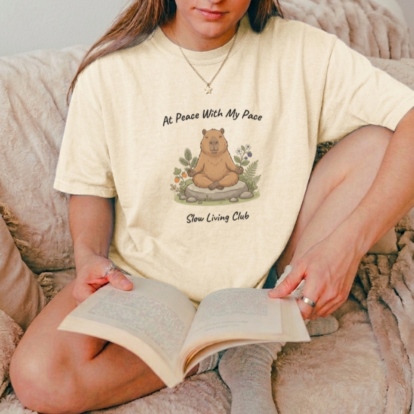 Woman wearing At Peace With My Pace meditating 
capybara graphic tee in Ivory reading a book 
on a cozy chair – Slow Living Club 
Comfort Colors shirt