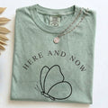 Close-up view of Comfort Colors sage green t-shirt with minimalist black butterfly line art and "HERE AND NOW" text, featuring a silver chain necklace.