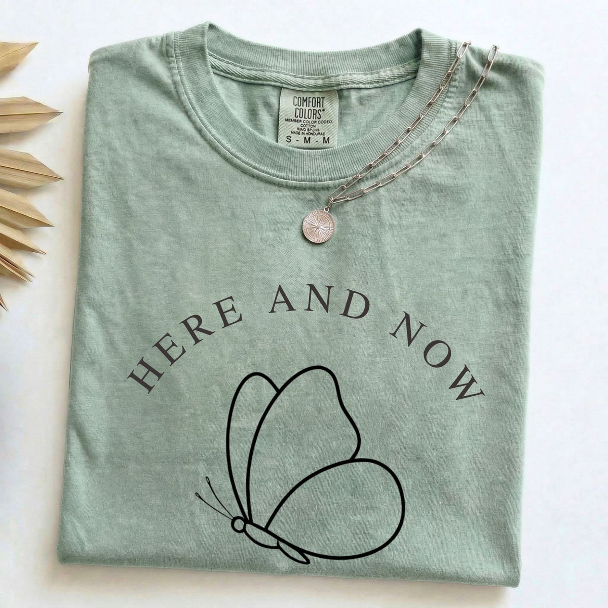 Close-up view of Comfort Colors sage green t-shirt with minimalist black butterfly line art and "HERE AND NOW" text, featuring a silver chain necklace.