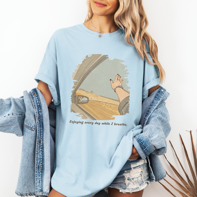 Light blue Comfort Colors graphic tee with a vintage-style open-road illustration seen from a car window and the phrase “Enjoying every day while I breathe.”