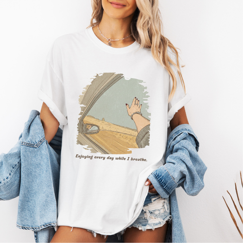 White Comfort Colors graphic tee with a vintage-style open-road illustration seen from a car window and the phrase “Enjoying every day while I breathe.”