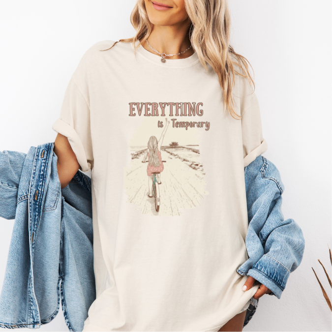 Ivory Comfort Colors graphic tee with a vintage-style illustration of a woman riding a bicycle down an open path and the phrase “Everything is Temporary.”