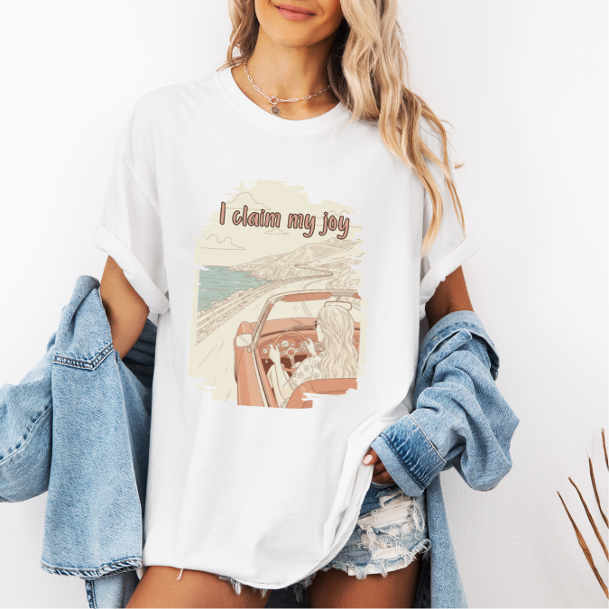 White Comfort Colors graphic tee with a vintage-style coastal driving illustration and the phrase “I claim my joy.”