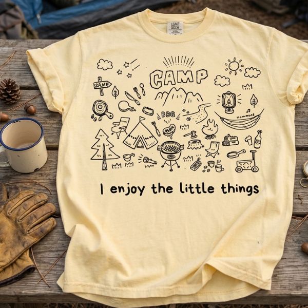 Butter yellow Comfort Colors t-shirt with camp doodle illustration and I enjoy the little things text, laid flat on a wooden surface surrounded by camping gloves, a metal mug, pinecones and acorns