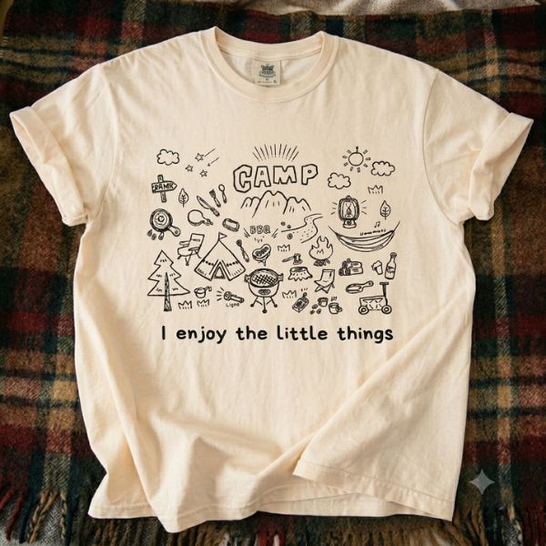 Ivory Comfort Colors t-shirt with hand-drawn camp doodle illustration and I enjoy the little things text, laid flat on a plaid flannel blanket