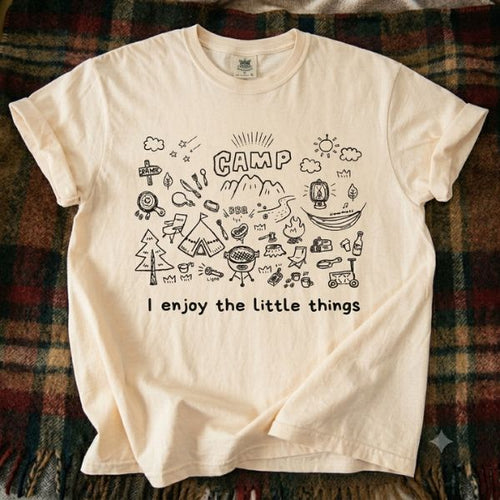 Ivory Comfort Colors t-shirt with hand-drawn camp doodle illustration and I enjoy the little things text, laid flat on a plaid flannel blanket