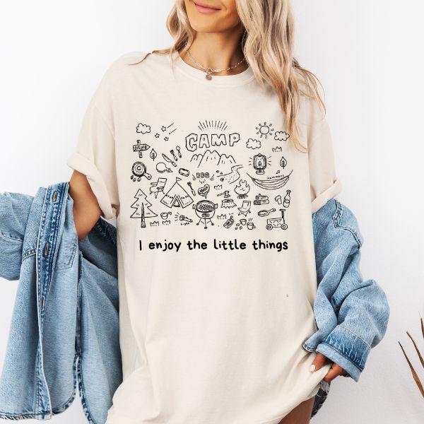 Woman wearing a ivory Comfort Colors t-shirt with a hand-drawn camp doodle illustration and the text I enjoy the little things, styled with a denim jacket
