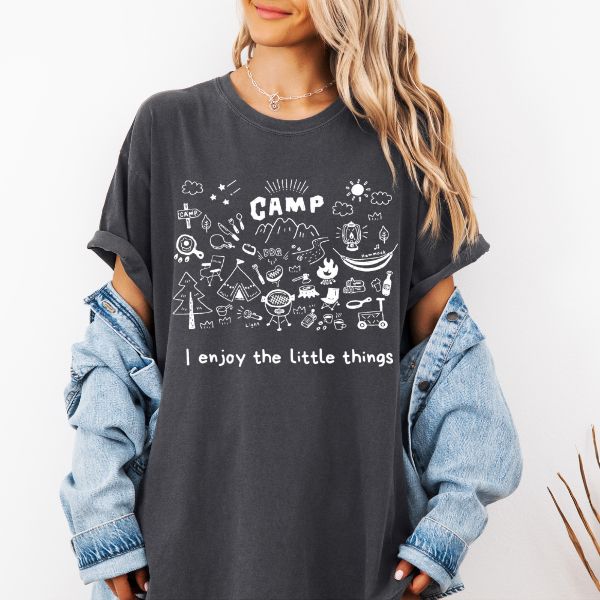 Woman wearing a dark pepper Comfort Colors t-shirt with white camp doodle illustration and I enjoy the little things text, styled with a denim jacket