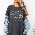 Woman wearing a dark pepper Comfort Colors t-shirt with white camp doodle illustration and I enjoy the little things text, styled with a denim jacket