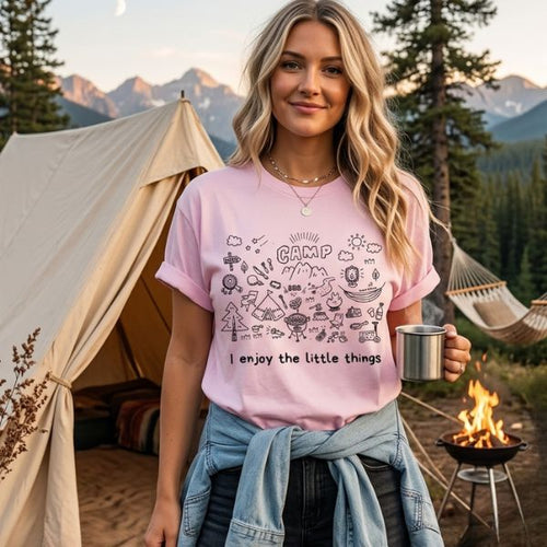 Woman wearing a Blossom Comfort Colors t-shirt with camp doodle illustration and I enjoy the little things text, standing in front of a canvas tent with pine trees, mountains, a hammock and a campfire in the background