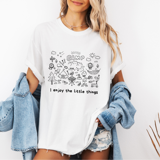 White Comfort Colors graphic tee with a blue camp doodle illustration featuring outdoor icons and the phrase “I enjoy the little things”
