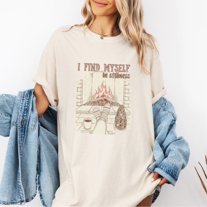 Ivory Comfort Colors graphic tee with a cozy fireplace illustration, coffee cup, socks, and the phrase “I find myself in stillness.”