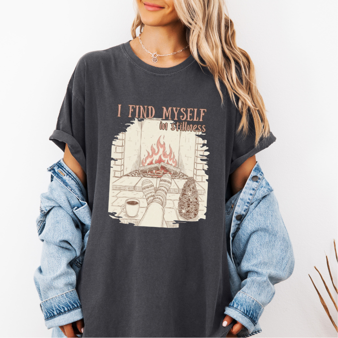 Pepper Comfort Colors graphic tee with a cozy fireplace illustration, coffee cup, socks, and the phrase “I find myself in stillness.”