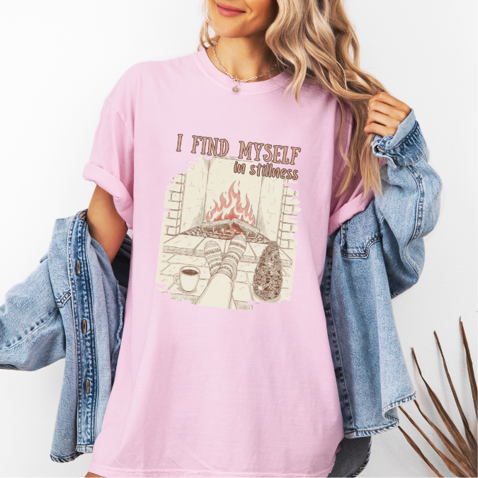 Pink Comfort Colors graphic tee with a cozy fireplace illustration, coffee cup, socks, and the phrase “I find myself in stillness.”