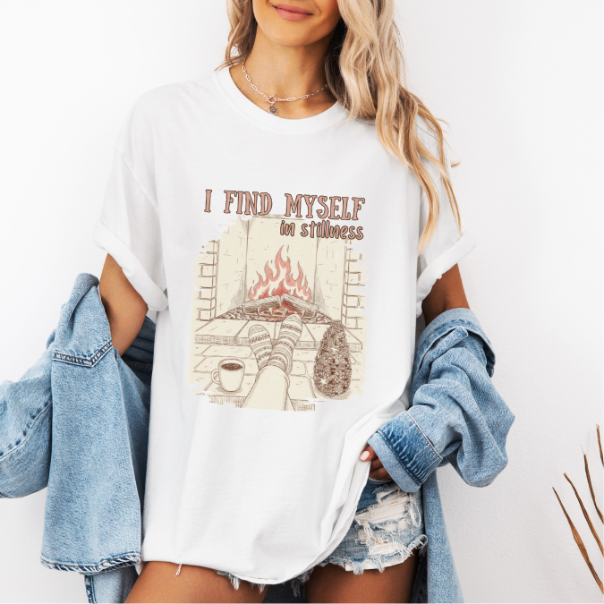 White Comfort Colors graphic tee with a cozy fireplace illustration, coffee cup, socks, and the phrase “I find myself in stillness.”