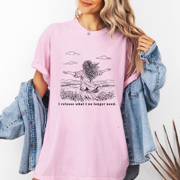 Woman wearing a pink Comfort Colors t-shirt with I release what I no longer need line art of a woman in a field with arms stretched wide under a cloudy sky, worn with a denim jacket