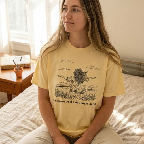 Woman wearing a butter yellow Comfort Colors t-shirt with I release what I no longer need field illustration, sitting on a bed in a warm sunlit bedroom with books and a small plant on a wooden nightstand