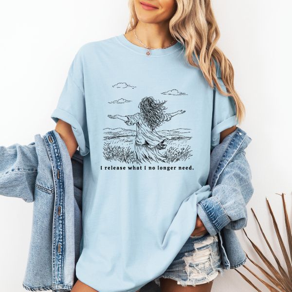 Woman wearing a light blue Comfort Colors t-shirt with I release what I no longer need line art of a woman in a field with arms stretched wide under a cloudy sky, worn with a denim jacket