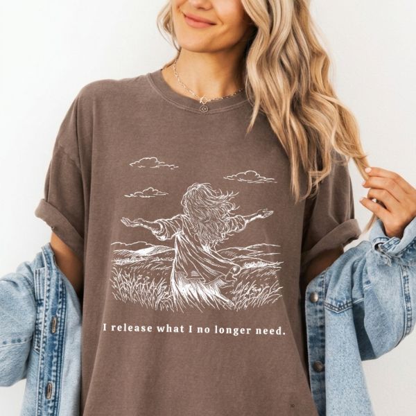 Woman wearing a espresso Comfort Colors t-shirt with white I release what I no longer need line art of a woman standing in a field with arms wide open, paired with a denim jacket