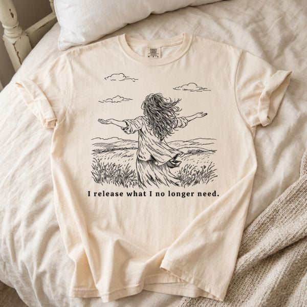 Ivory Comfort Colors t-shirt with I release what I no longer need line art illustration of a woman in a field with arms open, laid flat on a white bed