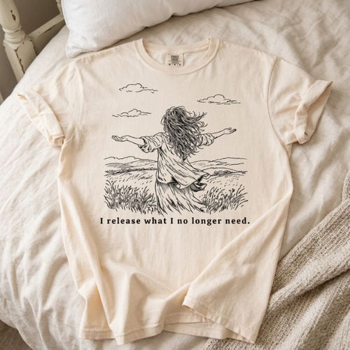 Ivory Comfort Colors t-shirt with I release what I no longer need line art illustration of a woman in a field with arms open, laid flat on a white bed