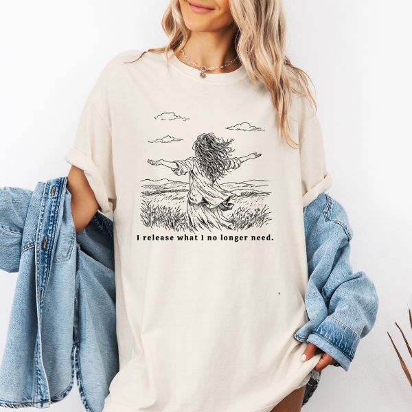 Woman wearing a IVORY Comfort Colors t-shirt with I release what I no longer need illustration of a woman with arms open in a windswept field, styled with a denim jacket