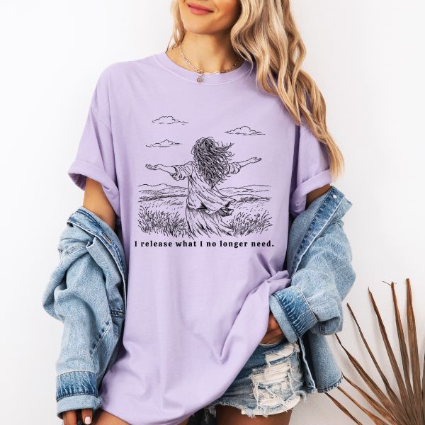 Woman wearing a light lavander Comfort Colors t-shirt with I release what I no longer need line art of a woman in a field with arms stretched wide under a cloudy sky, worn with a denim jacket