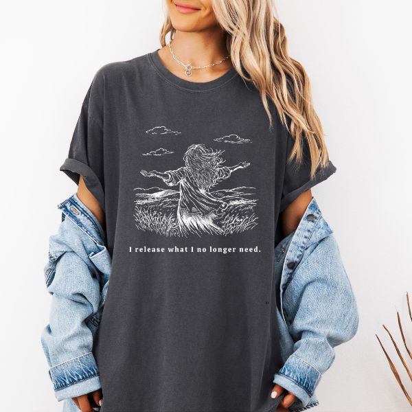 Woman wearing a dark pepper Comfort Colors t-shirt with white I release what I no longer need line art of a woman standing in a field with arms wide open, paired with a denim jacket