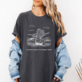 Woman wearing a dark pepper Comfort Colors t-shirt with white I release what I no longer need line art of a woman standing in a field with arms wide open, paired with a denim jacket