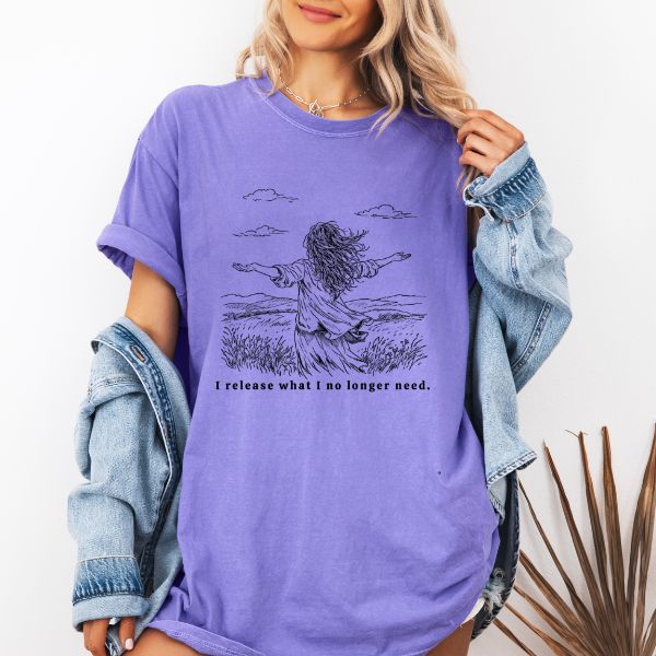 Woman wearing a lavander Comfort Colors t-shirt with I release what I no longer need line art of a woman in a field with arms stretched wide under a cloudy sky, worn with a denim jacket