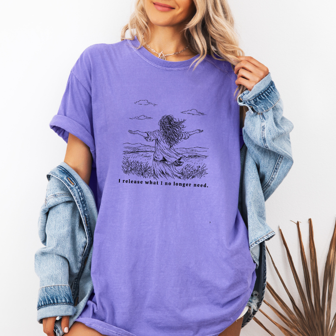 Violet Comfort Colors graphic tee with a line art illustration of a woman standing in an open field with arms open and the phrase “I release what I no longer need.”