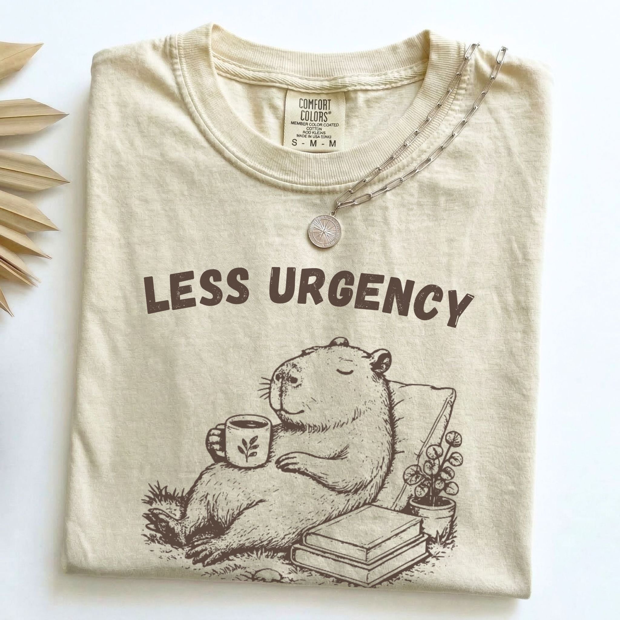 Less Urgency More Life capybara Comfort Colors tee in Ivory, folded flat lay with dried palm leaves