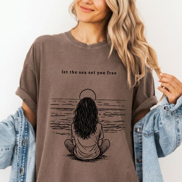 Woman wearing a Espresso Comfort Colors t-shirt with let the sea set you free ocean line art illustration of a woman in a meditative pose facing the sea, styled with a denim jacket