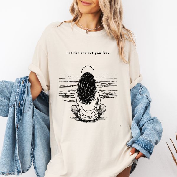 Woman wearing a Ivory Comfort Colors t-shirt with let the sea set you free line art of a woman sitting cross-legged facing the ocean under a sun halo, styled with a denim jacket