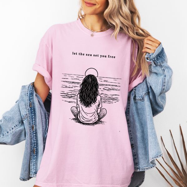Woman wearing a Blossom Comfort Colors t-shirt with let the sea set you free illustration of a woman sitting by the ocean with a sun halo, styled with a denim jacket