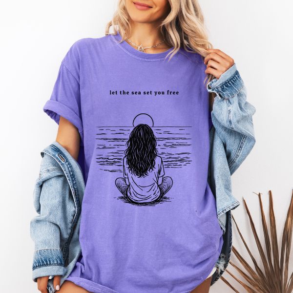 Woman wearing a Violet Comfort Colors t-shirt with let the sea set you free line art of a woman sitting meditatively facing the ocean under a sun halo, styled with a denim jacket