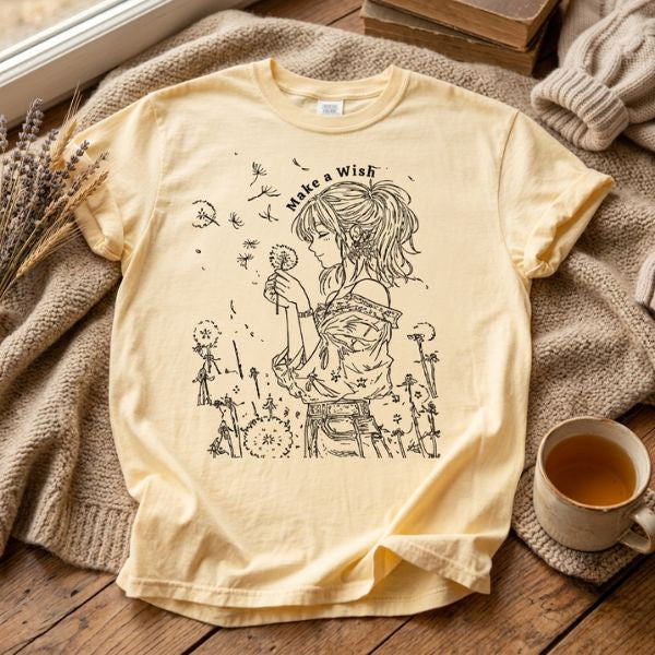 Butter yellow Comfort Colors t-shirt with Make a Wish dandelion girl illustration laid flat on a wooden surface next to a cozy knit sweater, lavender stems, and a ceramic mug of tea