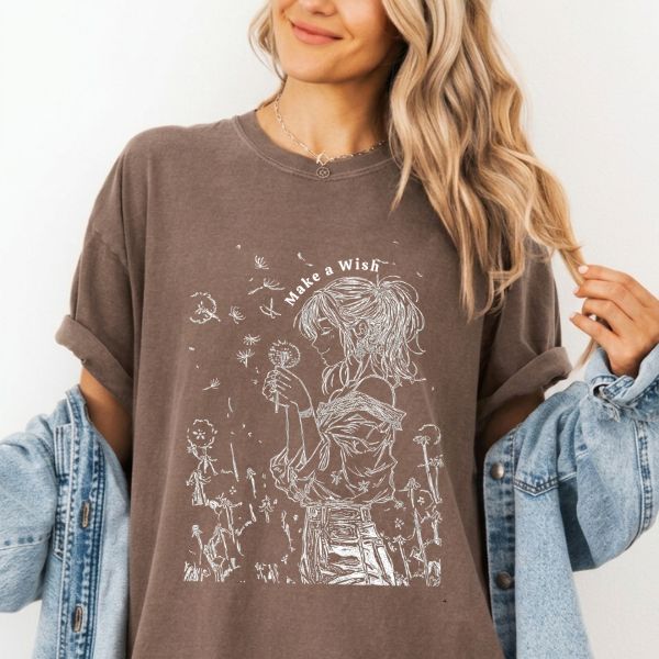 Close-up of a espresso Comfort Colors t-shirt featuring a white dandelion girl line art illustration with the text Make a Wish, worn by a woman with a denim jacket