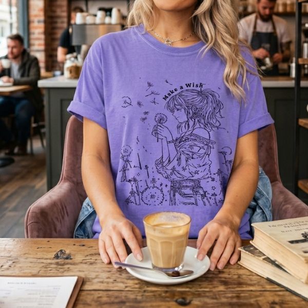 Woman wearing a violet Comfort Colors t-shirt with Make a Wish dandelion girl line art, sitting at a coffee shop table with a latte and vintage books nearby