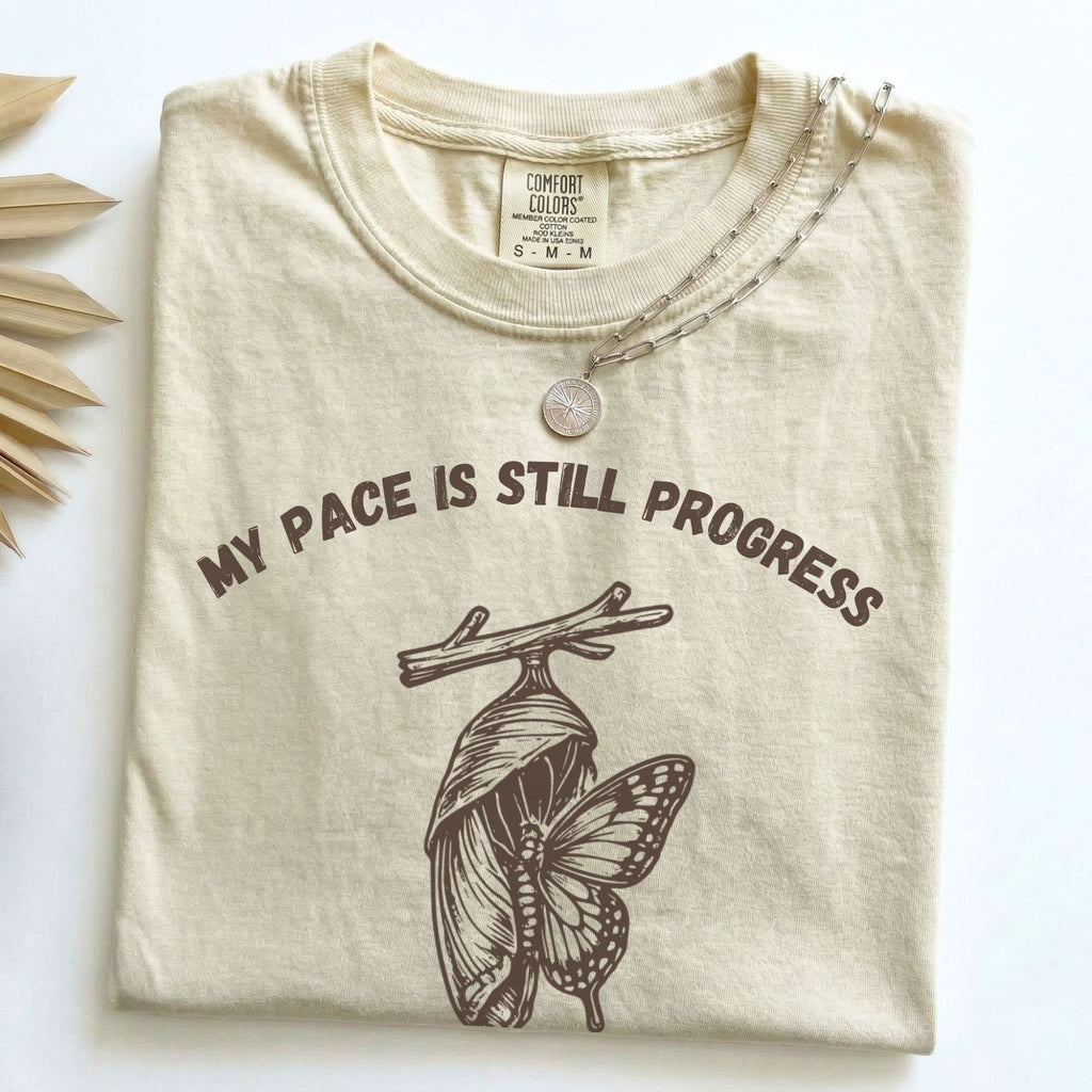 My Pace Is Still Progress chrysalis Comfort Colors tee in Ivory, folded flat lay with dried palm leaves